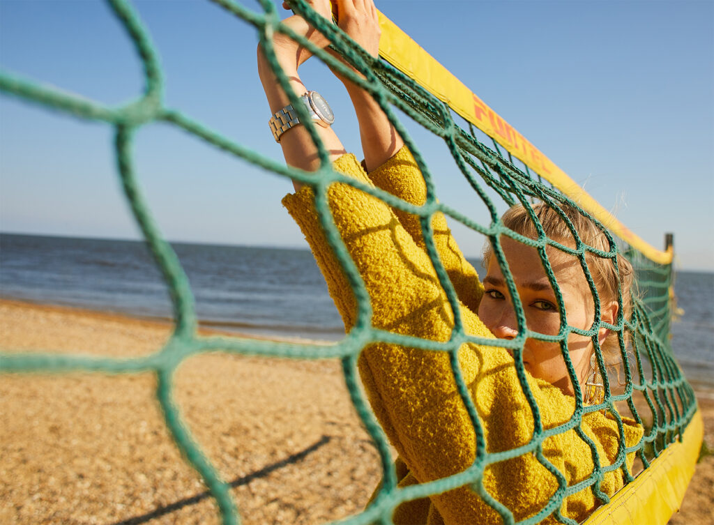Close-up portrait of a woman wearing a bright yellow sweater, looking forward from behind a green volleyball net on a sunny beach with the ocean in the background.
