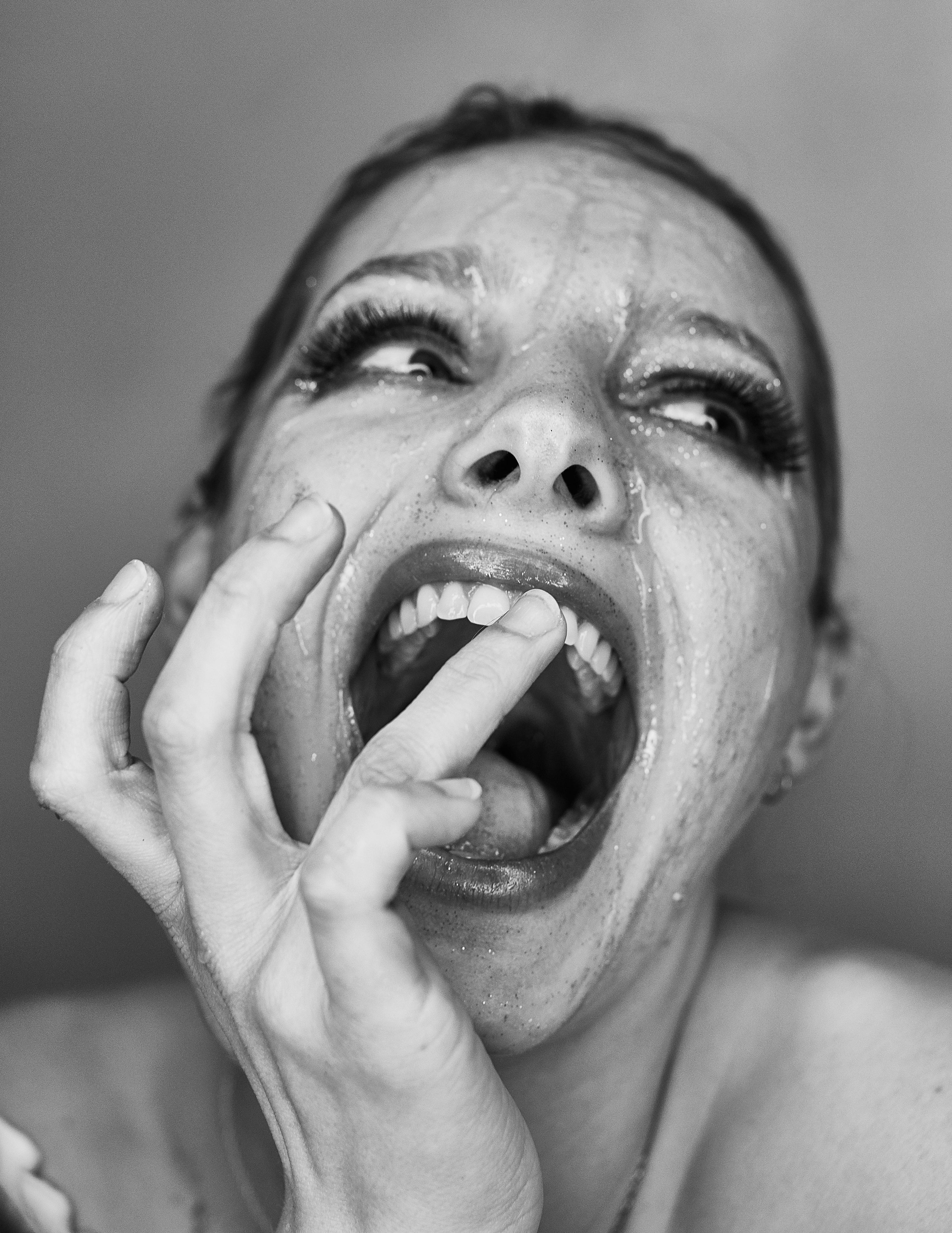 Close-up black and white portrait of a woman with her mouth wide open in a dramatic expression, a finger touching her teeth, with Honey glistening on her skin and face against a plain background.